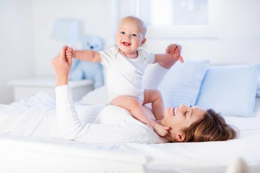 mastering playtime - mom laying on the bed with baby sitting on her stomach, both laughing and playing together