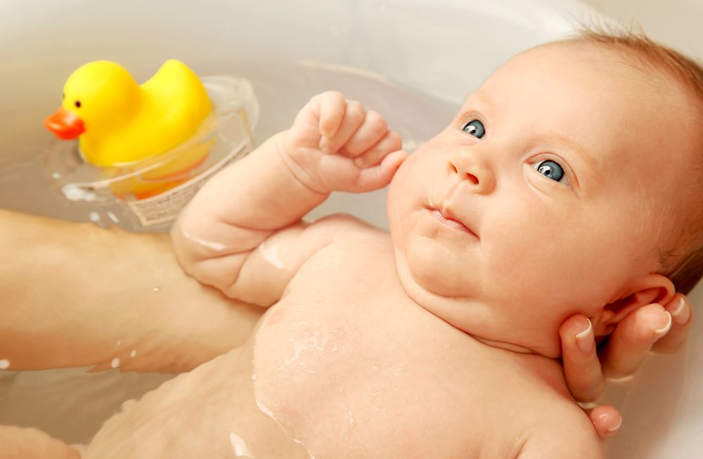 mom in bath with baby, holding baby in her arms while he looks up at her, rubber duckie in water floating next to them