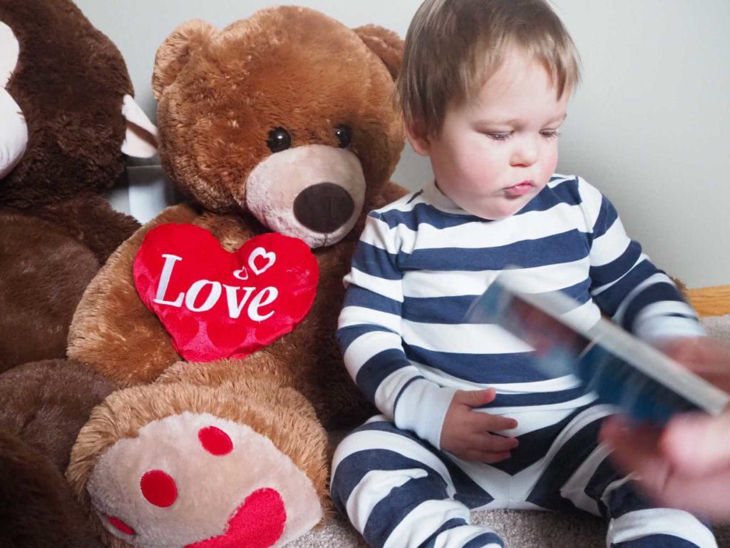 baby in Peejamas reading a book and sitting next to giant teddy bear