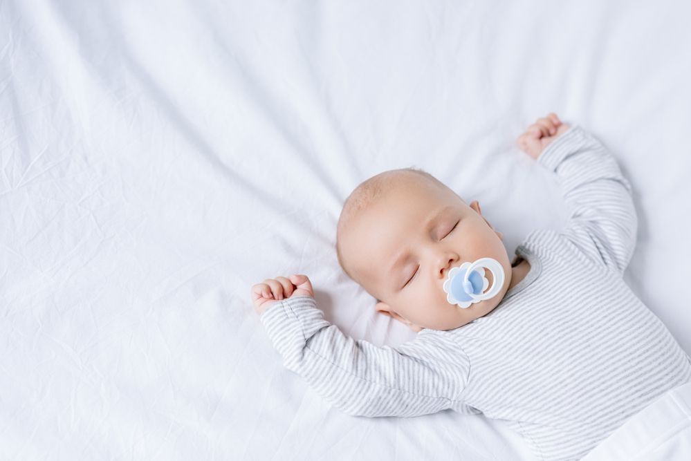 baby sleeping on the bed with arms up and pacifier in his mouth