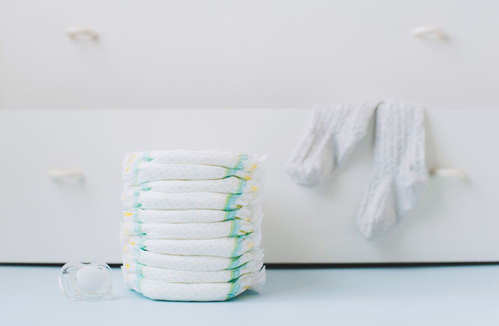 A stack of diapers on the background of a white dresser with blankets hanging over the bottom dresser drawer