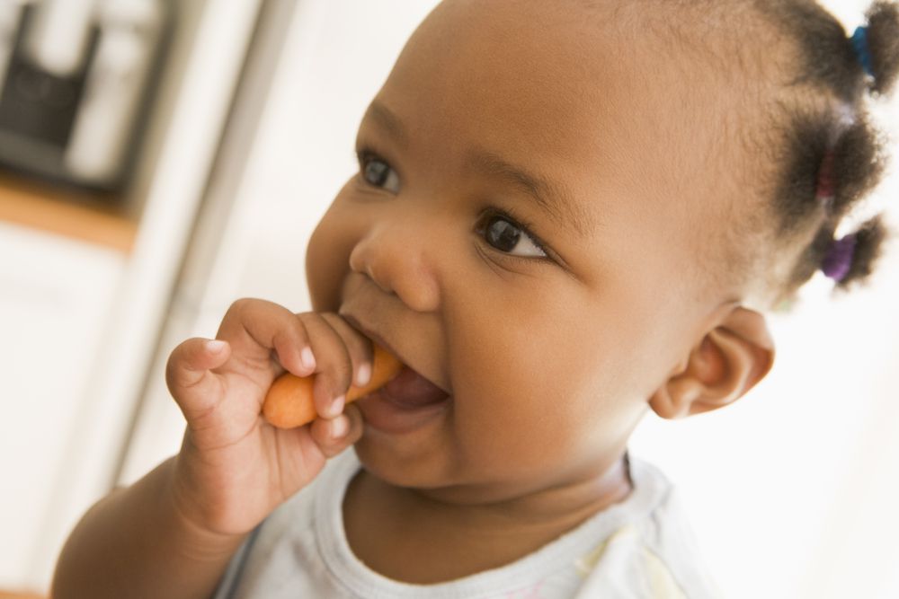Baby eating a carrot. When babies eat more solid foods, it's normal to drink less which leads to normal bottle refusal.