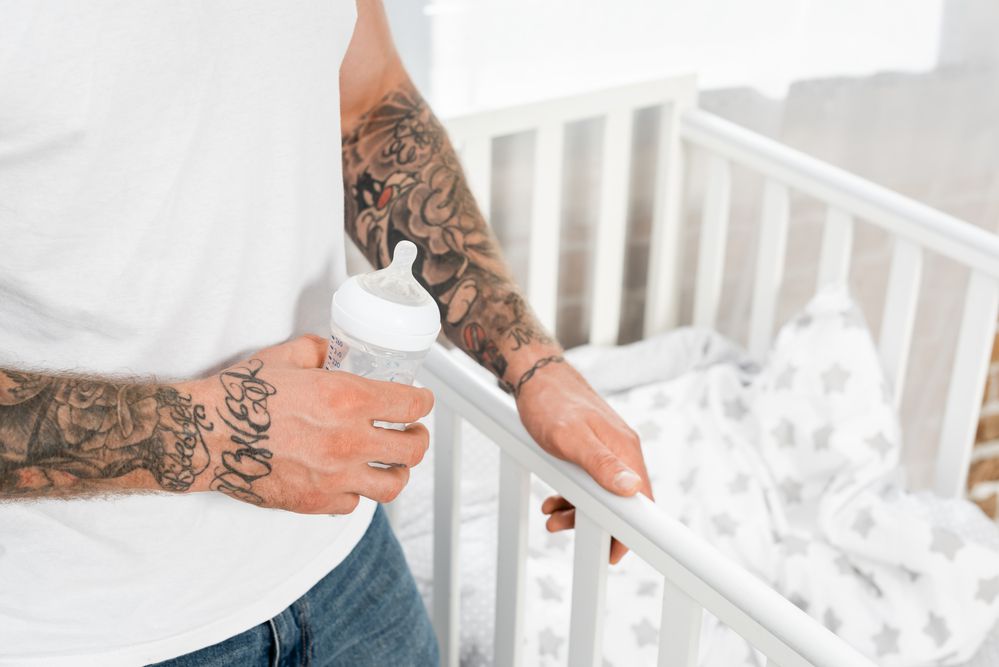 Dad with tattooed arms holding a bottle standing by a crib
