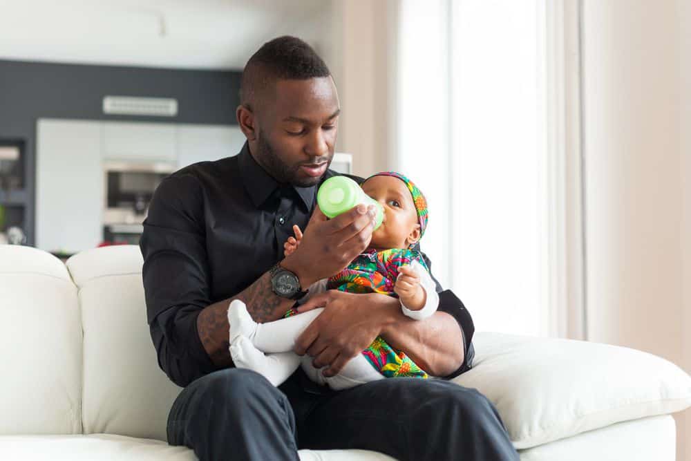 african american father sitting on the couch and using paced bottle feeding to feed his baby